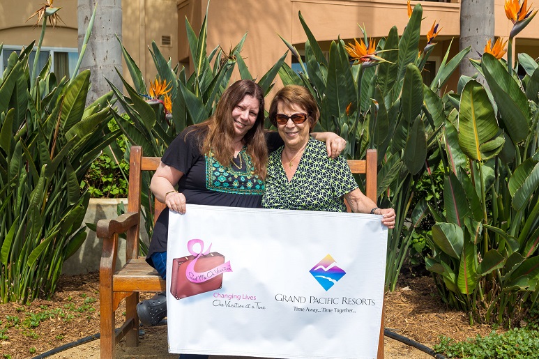 Gisele, a Send Me On Vacation Survivor, sits in the garden at Grand Pacific Palisades with her mother while holding a sign with the Grand Pacific Resorts and Send Me On Vacation logos